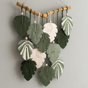 May include: A macrame wall hanging with a wooden branch and green and white leaves. The leaves are made of yarn and are hanging from the branch.