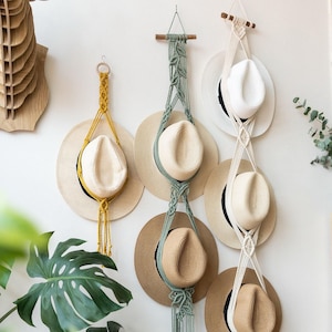 May include: A collection of straw hats displayed on macrame hangers against a white wall. The hats have various brim styles and colors, including natural, white, and tan. The macrame hangers are in yellow, sage green, and cream colors.
