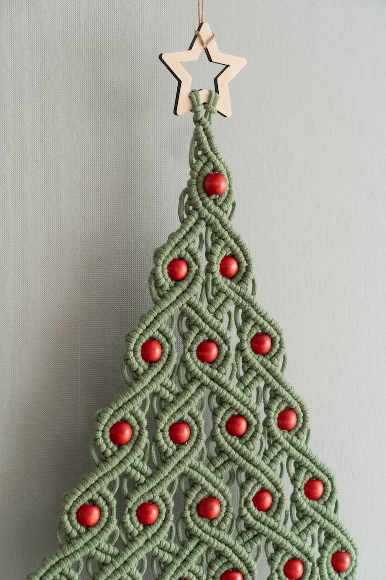 K&ouml;nnte beinhalten: Ein gr&uuml;ner Makramee-Weihnachtsbaum-Wandbehang mit roten Holzperlen und einem Holzstern an der Spitze.