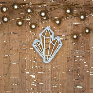 May include: White plastic cookie cutter shaped like a crystal. The cutter is on a cork background with gold flecks.