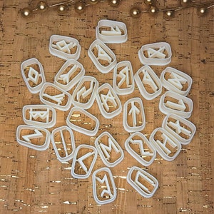 May include: A set of white plastic cookie cutters shaped like different runes. The cutters are scattered on a cork surface.