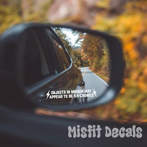 May include: A black vehicle side mirror reflecting a road lined with autumn trees. A white vinyl decal is applied to the mirror glass, featuring two lightning bolt icons and the text: OBJECTS IN MIRROR MAY APPEAR TO BE KA-CHOWED.