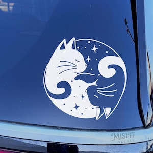 May include: A white vinyl decal applied to a dark car window, featuring a circular design of a cat and a bird facing each other. The cat is on the left and the bird is on the right, surrounded by small four-pointed stars within the circle.
