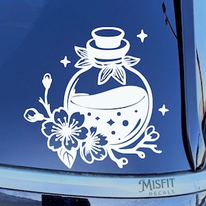 May include: A white vinyl decal featuring a round potion bottle with a cork stopper, surrounded by cherry blossom flowers and small four-pointed stars. The text MISFIT DECALS is printed in a small font at the bottom right of the design.