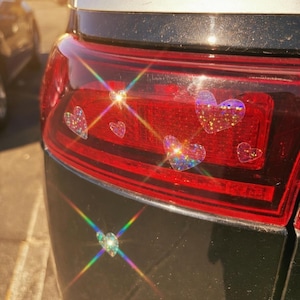 May include: Close-up of a car's taillight with several holographic heart-shaped stickers. The stickers are iridescent, reflecting a rainbow of colors. The taillight is red and black, with a shiny surface.