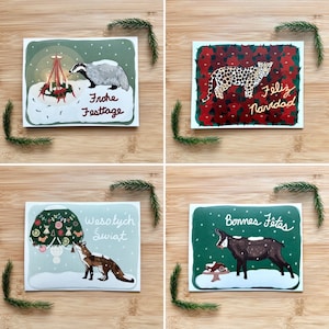 May include: Four Christmas cards featuring different animals and holiday greetings. The cards are on a wooden background. The first card features a badger with the text "Frohe Festtage". The second card features a jaguar with the text "Feliz Navidad". The third card features a fox with the text "Wesołych Świąt". The fourth card features a mountain goat with the text "Bonnes Fêtes".