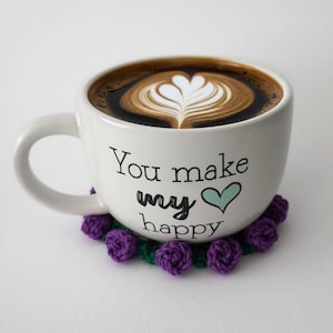 Könnte beinhalten: Eine weiße Keramiktasse gefüllt mit Kaffee, mit Latte Art in Herzform. Die Tasse trägt den Text "You make my happy" und ein türkisfarbenes Herz. Sie steht auf einem lila Häkeluntersetzer mit grünem Rand.