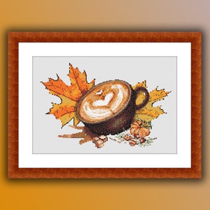Könnte beinhalten: Eine braune Keramiktasse gefüllt mit einem cremigen Latte-Kunstdesign, umgeben von Herbstblättern in Orange- und Gelbtönen. Die Tasse steht auf einer weißen Oberfläche mit verstreuten Nüssen.