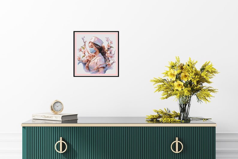 May include: A framed painting of a woman wearing a white nurse's uniform and a blue face mask. She is surrounded by pink flowers. The painting is on a white wall above a green dresser with gold handles.