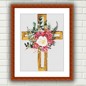 Puede incluir: Una cruz dorada con un arreglo floral de rosas rosas y blancas, vegetación y ramitas. La cruz está enmarcada en un marco de madera marrón.