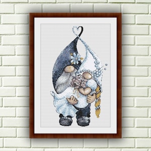 May include: A cross-stitch pattern depicting a bride and groom gnome couple holding hands. The bride is wearing a white dress with blue flowers and a veil. The groom is wearing a blue hat and a black suit. The couple are standing in front of a heart-shaped design.