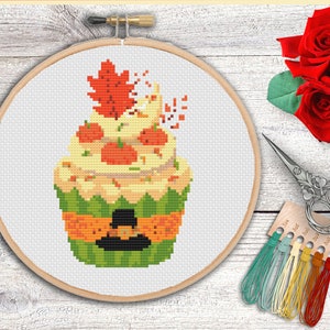 May include: A cross-stitch pattern of a fall-themed cupcake with orange and yellow pumpkins, a brown hat, and a red and orange maple leaf on top. The cupcake is on a green base with an orange and yellow band.
