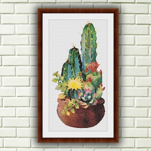 May include: A framed cross-stitch pattern of a potted cactus plant with green, yellow, and red flowers. The cactus is in a brown pot with a white background.