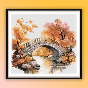 May include: Cross-stitch artwork depicting a stone bridge over a calm river, with trees displaying orange and yellow autumn foliage. The artwork is framed in a black square frame.