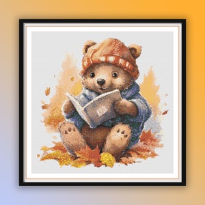 May include: A brown teddy bear wearing a red and white hat and a blue jacket sits on a pile of autumn leaves and reads a book.