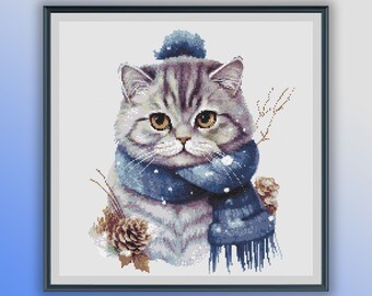 Puede incluir: Un gato atigrado gris con una bufanda azul y un gorro azul con pompón. El gato mira al espectador con una expresión seria. El fondo es blanco.