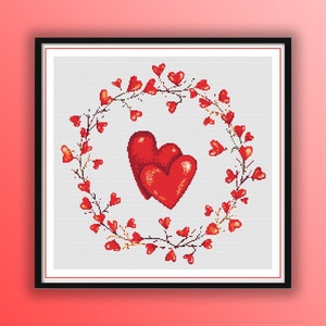 May include: A framed cross-stitch design featuring two large red hearts at the center, surrounded by a wreath of red heart-shaped leaves and branches. The background is a light gray color.