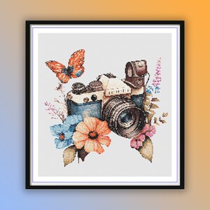 Op de afbeelding: Een vintage camera met een blauwe lens omringd door aquarelbloemen, bladeren en een vlinder. De camera is wit met bruine accenten en een zwarte band.