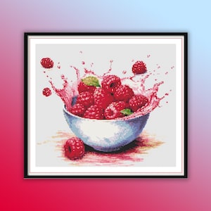 Puede incluir: Un tazón blanco lleno de frambuesas rojas, con un chorro de líquido rosa alrededor del tazón. Las frambuesas están dispuestas en un racimo, con algunas que se caen del tazón.