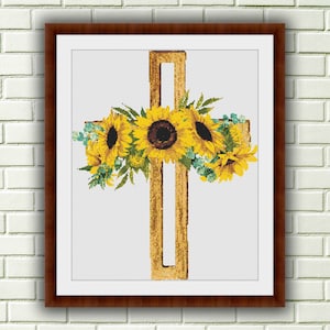 Puede incluir: Una cruz de madera con un arreglo floral de girasoles y vegetación. Los girasoles son amarillos con centros oscuros y la vegetación es de un verde claro.