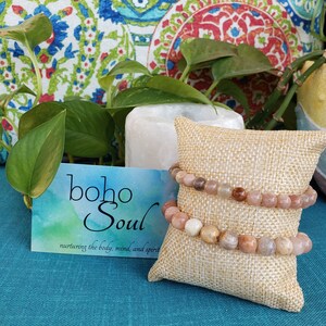 May include: Two beaded bracelets with neutral-toned stones rest on a small burlap pillow. A business card with the text "boho Soul" and the tagline "nurturing the body, mind, and spirit" is visible. The background includes a patterned pillow and greenery.