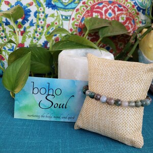 May include: A beaded bracelet with green, brown, and gray stones rests on a small burlap pillow. A card with the words "boho Soul" and the phrase "nurturing the body, mind, and spirit" is in the foreground. The background includes a white candle and decorative pillows.