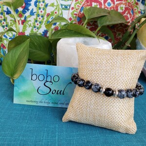 May include: A black and gray beaded bracelet with silver accents rests on a burlap pillow. The bracelet is displayed in front of a card that reads "boho Soul" with the tagline "nurturing the body, mind, and spirit."