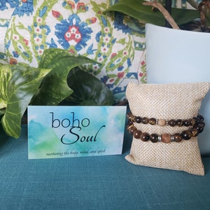 May include: Two beaded bracelets with brown and tan stones rest on a burlap pillow. A business card with the text "boho Soul" and the phrase "nurturing the body, mind, and spirit" is in front of the bracelets. Green plants and a blue pot are in the background.