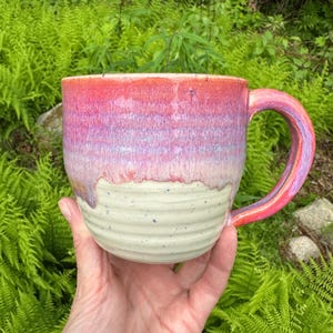 May include: A handmade ceramic mug with a vibrant color gradient. The top half transitions from red to purple, while the bottom is a creamy white with subtle speckles. The handle mirrors the color scheme. A unique piece for enjoying beverages.