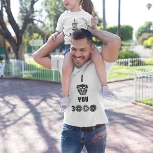 K&ouml;nnte beinhalten: Ein Vater und seine Tochter tragen passende wei&szlig;e T-Shirts mit einem schwarz-wei&szlig;en Motiv von Iron Mans Arc-Reaktor und dem Text "Ich liebe dich 3000". Der Vater tr&auml;gt seine Tochter auf seinen Schultern. Beide l&auml;cheln und schauen in die Kamera.