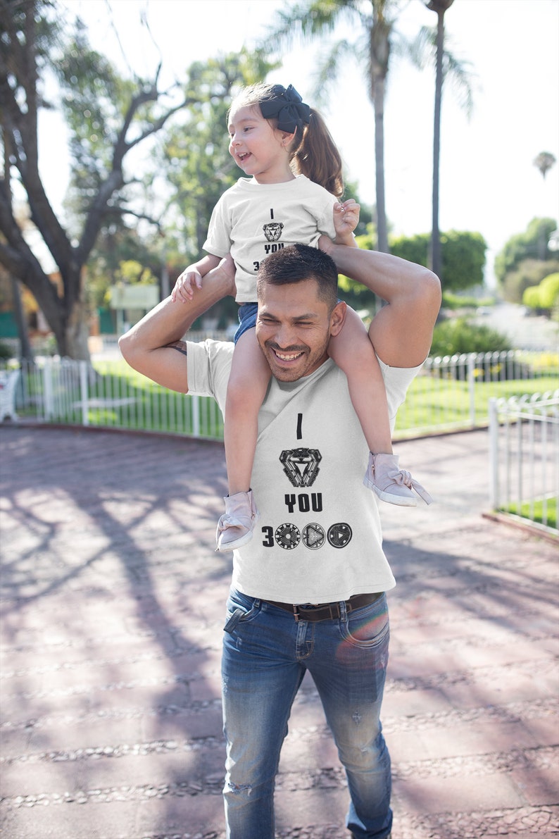 K&ouml;nnte beinhalten: Ein Vater und seine Tochter tragen passende wei&szlig;e T-Shirts mit einem schwarz-wei&szlig;en Motiv von Iron Mans Arc-Reaktor und dem Text "Ich liebe dich 3000". Der Vater tr&auml;gt seine Tochter auf seinen Schultern. Beide l&auml;cheln und schauen in die Kamera.