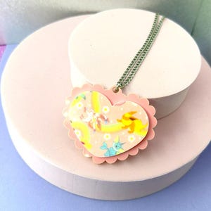 Puede incluir: Un collar con colgante en forma de corazón con borde festoneado rosa. El colgante presenta un fondo rosa claro con un diseño de dibujos animados colorido. La cadena del collar es de color plateado. El colgante se exhibe sobre un soporte cilíndrico blanco.