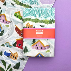 May include: A white tea towel with a winter village scene printed on it. The design features colorful houses, snow-covered trees, and a winding road. The tea towel has a pink label with the text "tickled pink" on it.
