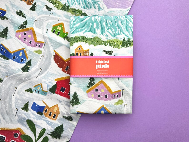 May include: A white tea towel with a winter village scene printed on it. The design features colorful houses, snow-covered trees, and a winding road. The tea towel has a pink label with the text "tickled pink" on it.