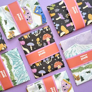 May include: A set of ten tea towels featuring various winter-themed designs. The towels are white with colorful illustrations of houses, mountains, and mushrooms. Each towel has a pink label with the text "Tikkled Pink" printed on it.