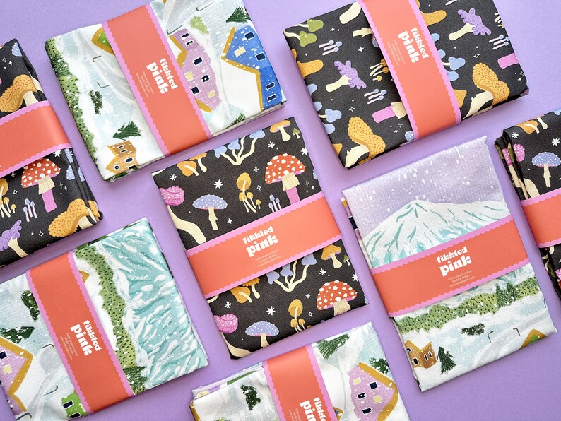 May include: A set of ten tea towels featuring various winter-themed designs. The towels are white with colorful illustrations of houses, mountains, and mushrooms. Each towel has a pink label with the text "Tikkled Pink" printed on it.