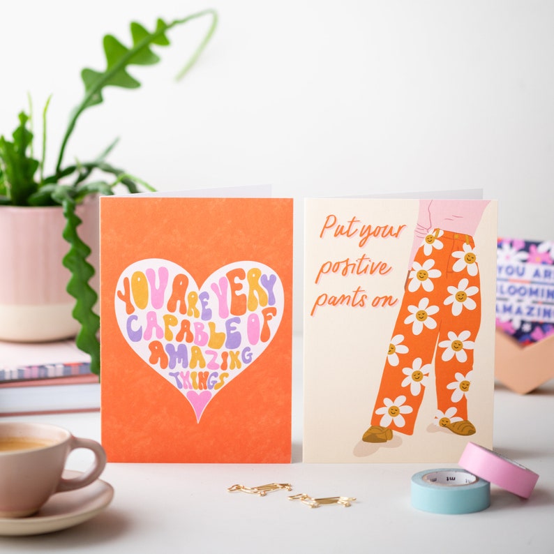 May include: Two greeting cards with positive messages. The first card has an orange background with a heart shape containing the text "You are very capable of amazing things". The second card has a cream background with a person wearing orange pants with a daisy pattern and the text "Put your positive pants on".