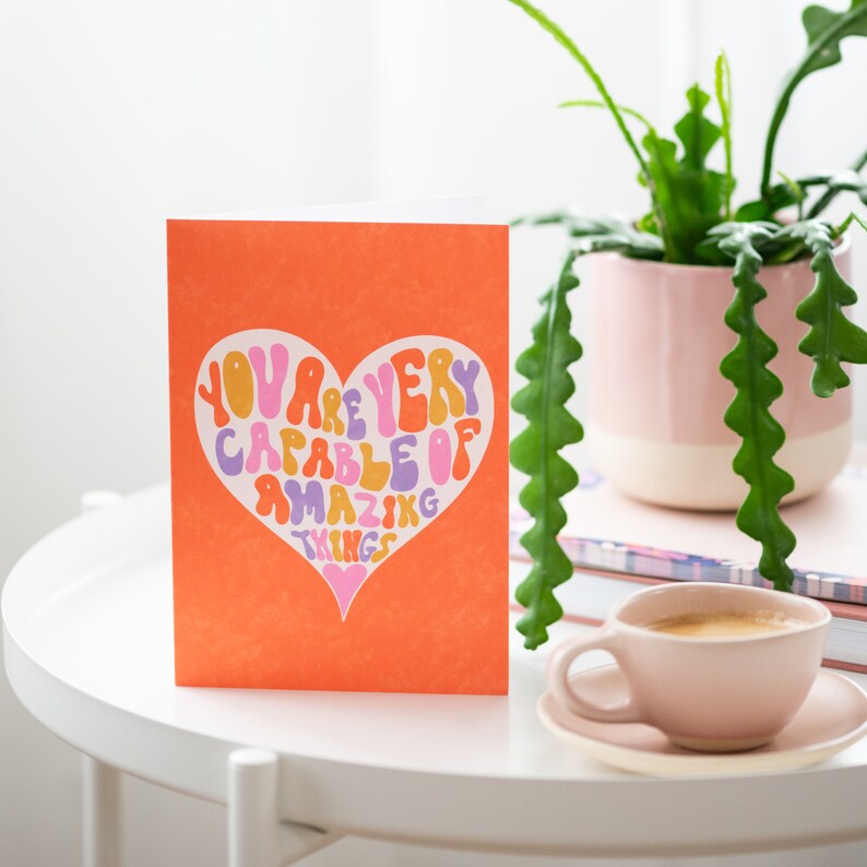 May include: An orange card with a heart-shaped design. The heart is filled with colorful text that reads "You are very capable of amazing things".