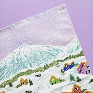 May include: A white tea towel with a winter scene printed on it. The scene features a snowy mountain range, a small village with houses, and a light purple sky with white stars.