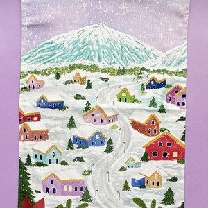 May include: A white kitchen towel with a winter scene of a snowy mountain and colorful houses. The houses are painted in shades of pink, blue, green, red, and yellow. The scene is set against a light purple sky with white snowflakes.
