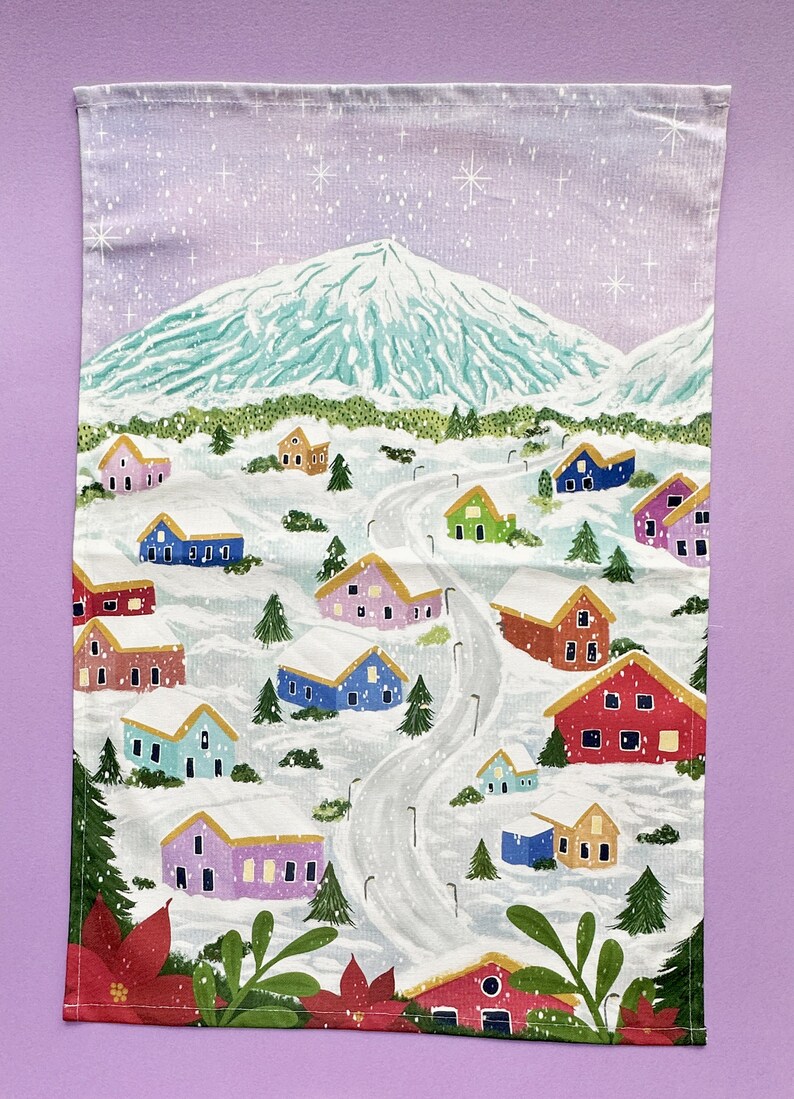 May include: A white kitchen towel with a winter scene of a snowy mountain and colorful houses. The houses are painted in shades of pink, blue, green, red, and yellow. The scene is set against a light purple sky with white snowflakes.