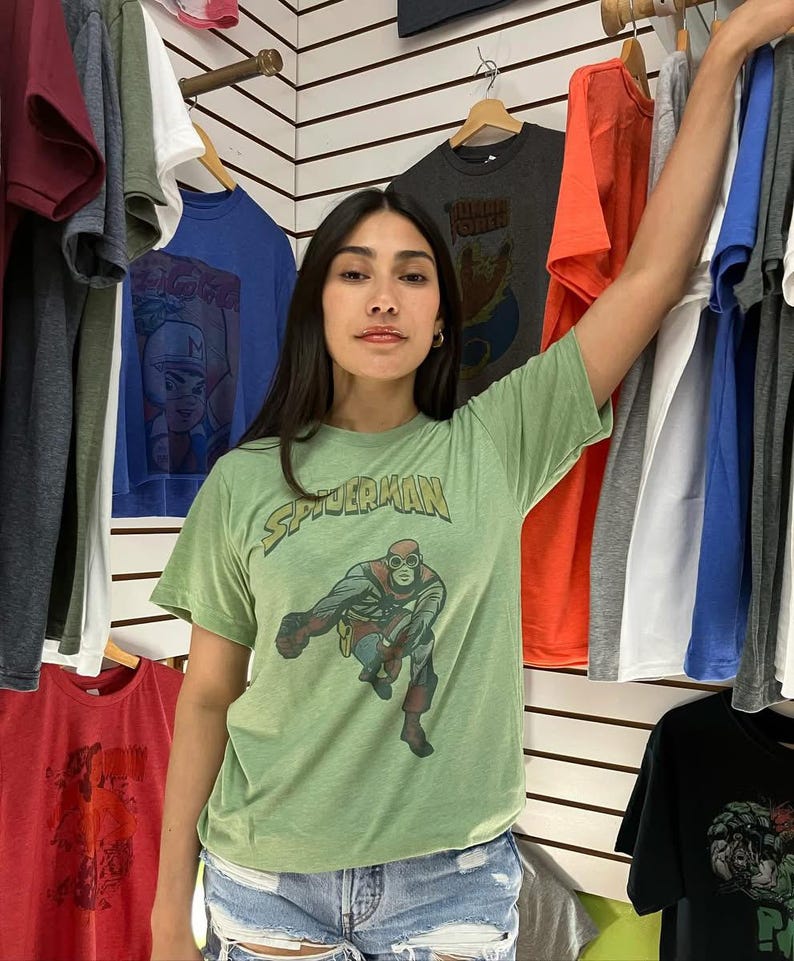 K&ouml;nnte beinhalten: Ein hellgr&uuml;nes T-Shirt mit einem Vintage-Spider-Man-Motiv. Das Bild zeigt Spider-Man in einer klassischen Pose, mit dem Wort "SPIDERMAN" dar&uuml;ber. Das T-Shirt ist von anderen T-Shirts auf Kleiderb&uuml;geln umgeben.