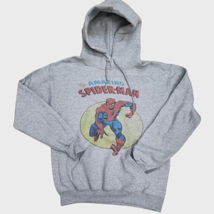 May include: A gray hooded sweatshirt featuring a graphic of Spider-Man in a yellow circle. The text "AMAZING SPIDER-MAN" is above the character. The sweatshirt has a drawstring hood and a front pocket.