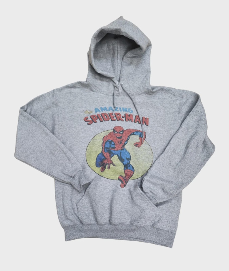 May include: A gray hooded sweatshirt featuring a graphic of Spider-Man in a yellow circle. The text "AMAZING SPIDER-MAN" is above the character. The sweatshirt has a drawstring hood and a front pocket.