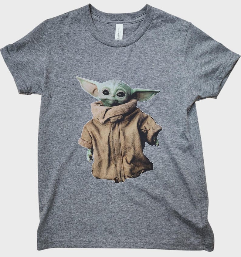 May include: A gray t-shirt featuring a graphic of a green alien character wearing a brown coat. The character has large ears and dark eyes. The t-shirt is made of a soft material.