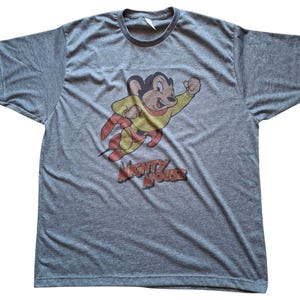 Puede incluir: Camiseta gris con una imagen de dibujos animados de Mighty Mouse con un traje de superhéroe amarillo y rojo. El texto "MIGHTY MOUSE" está impreso en rojo y marrón debajo del personaje. La camiseta tiene cuello redondo y mangas cortas.