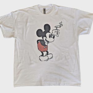 May include: White t-shirt featuring a graphic of Mickey Mouse smoking. The cartoon character is depicted in black, red, and white, holding a smoking object. The shirt is a crew neck style.