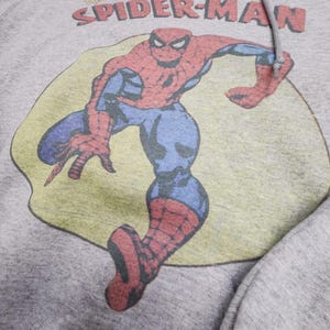 May include: A gray hooded sweatshirt featuring a vintage graphic of Spider-Man. The superhero is depicted in his classic red and blue suit, set against a yellow and green background. The words "AMAZING SPIDER-MAN" are printed above the image in red and blue.
