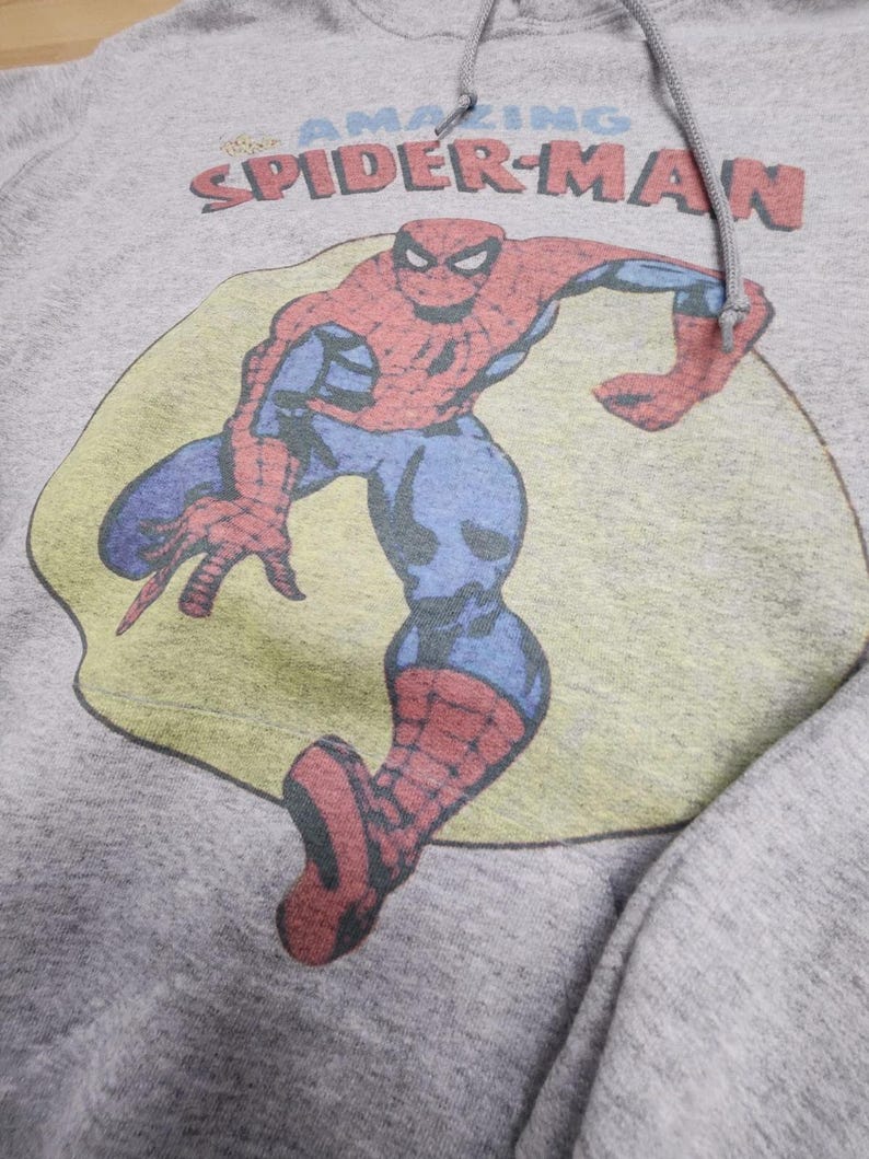 May include: A gray hooded sweatshirt featuring a vintage graphic of Spider-Man. The superhero is depicted in his classic red and blue suit, set against a yellow and green background. The words "AMAZING SPIDER-MAN" are printed above the image in red and blue.