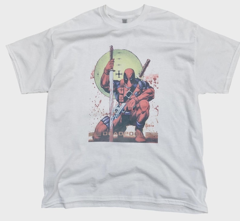 May include: White t-shirt featuring a comic book illustration of Deadpool kneeling, holding a sword, with a green target background and the word "DEADPOOL" at the bottom. The design includes red and brown tones.